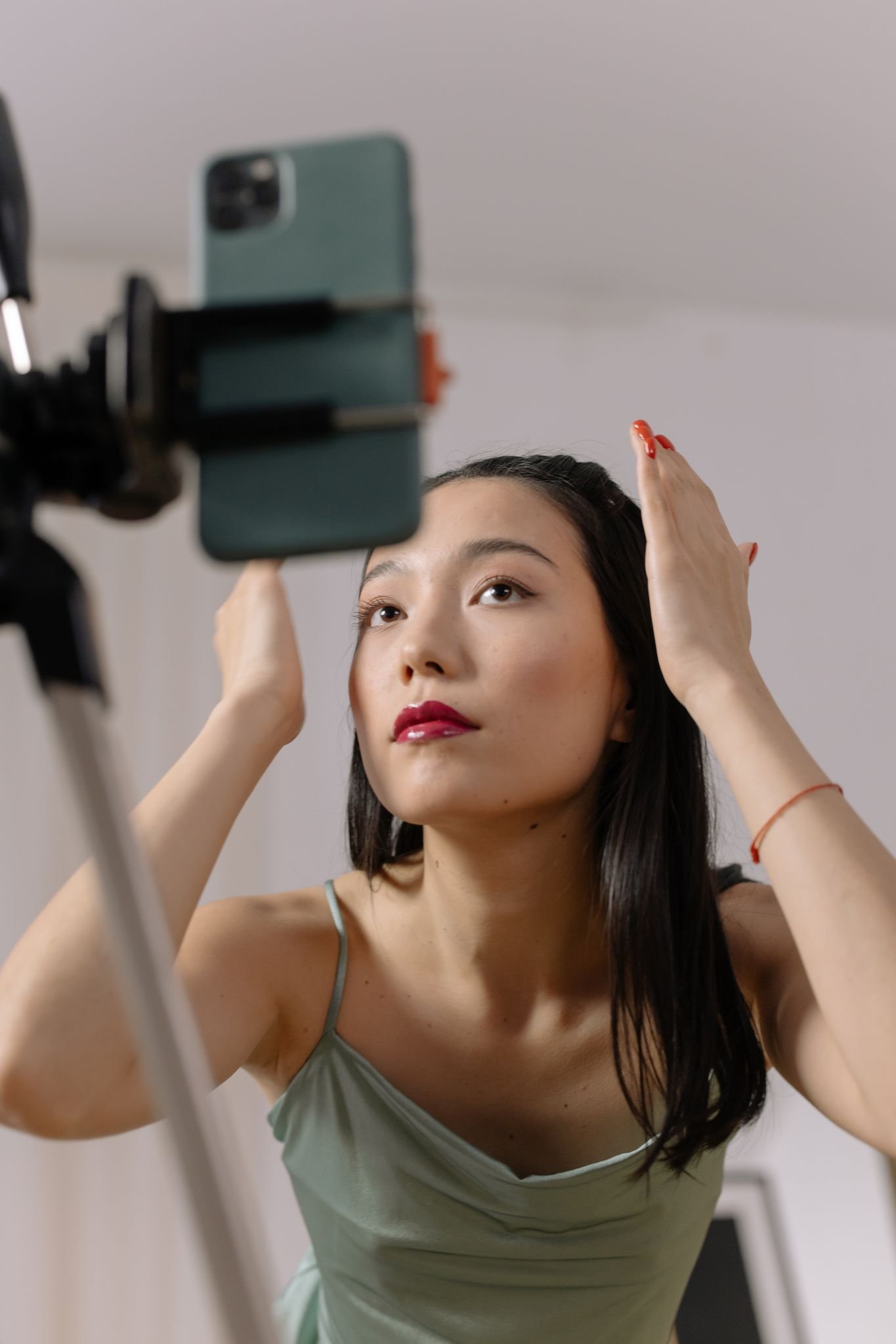 Asian woman adjusts hair and makeup in front of smartphone for live online content creation.