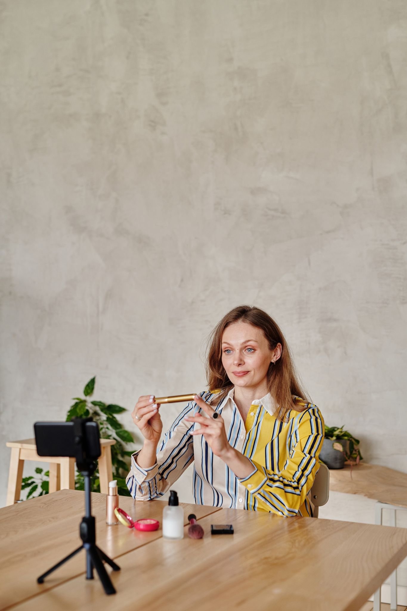 Caucasian woman live streaming a beauty vlog, showcasing makeup products on a wooden table.